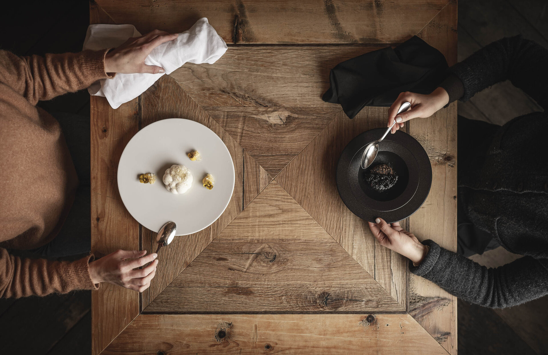 Zwei Personen sitzen an einem alten Holztisch und essen monochrome Gerichte - weißes Gericht auf weißem Teller und schwarzes Gericht auf schwarzem Teller