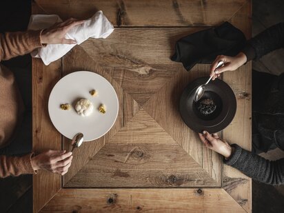 Zwei Personen sitzen an einem alten Holztisch und essen monochrome Gerichte - weißes Gericht auf weißem Teller und schwarzes Gericht auf schwarzem Teller