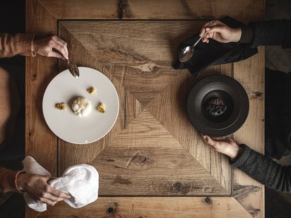 Zwei Personen sitzen an einem alten Holztisch und essen monochrome Gerichte - weißes Gericht auf weißem Teller und schwarzes Gericht auf schwarzem Teller