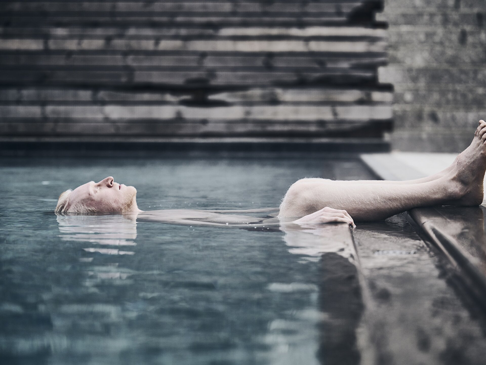 Person mit weißer Haut und weißem Haar lässt sich im Pool treiben, dabei liegen die Beine am Beckenrand auf