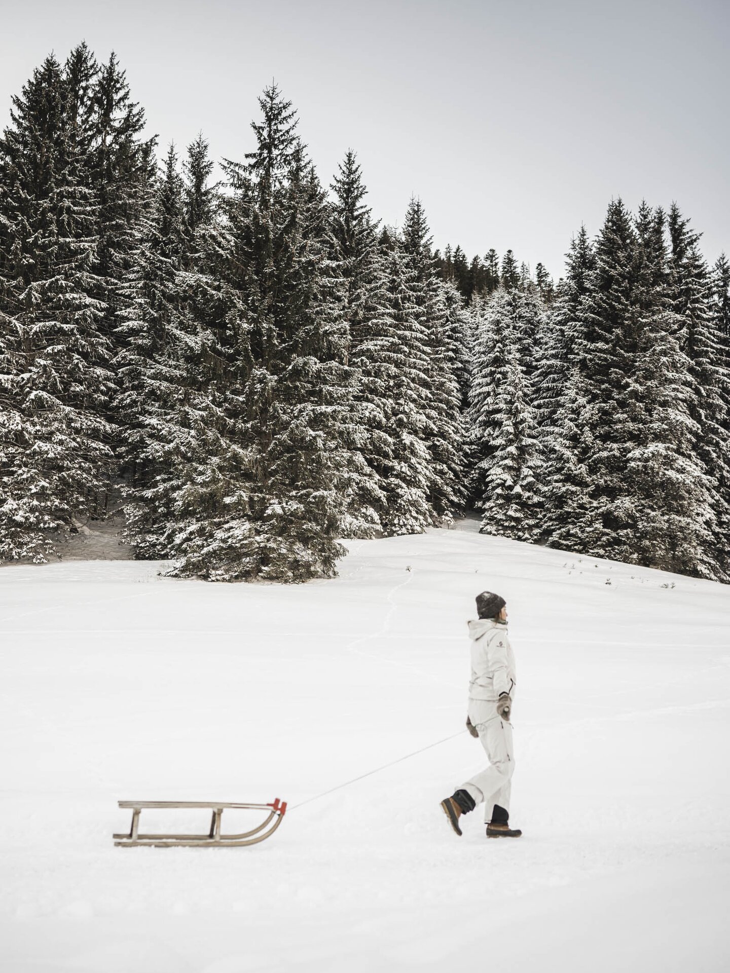 Person im beigen Winteroutfit zieht eine Rodel im verschneiten Wald hinter sich her