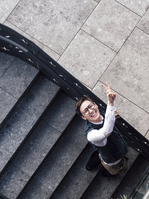 Toni Schlachter - Hoteldirektor - steht auf der Treppe im Außenbereich des Wellnesshotels Der Engel und lächelt in die Kamera und zeigt Peace
